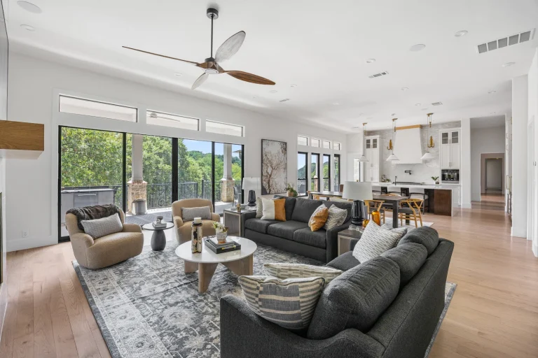 A spacious open concept living room with a large gray sectional sofa, tan armchairs, and a round white coffee table on a patterned rug featuring a modern wood ceiling fan and a view of a lush green landscape through sliding glass doors. Hero